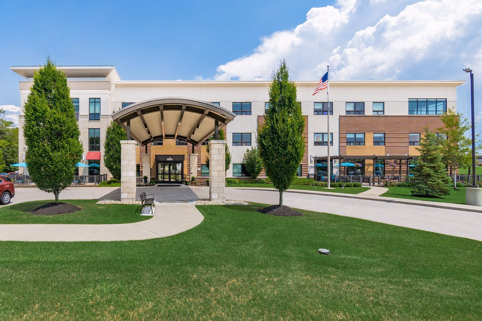 Front exterior view of a three-story senior living facility building with a covered entrance, green lawn, trees, an American flag, and a clear blue sky with some clouds.