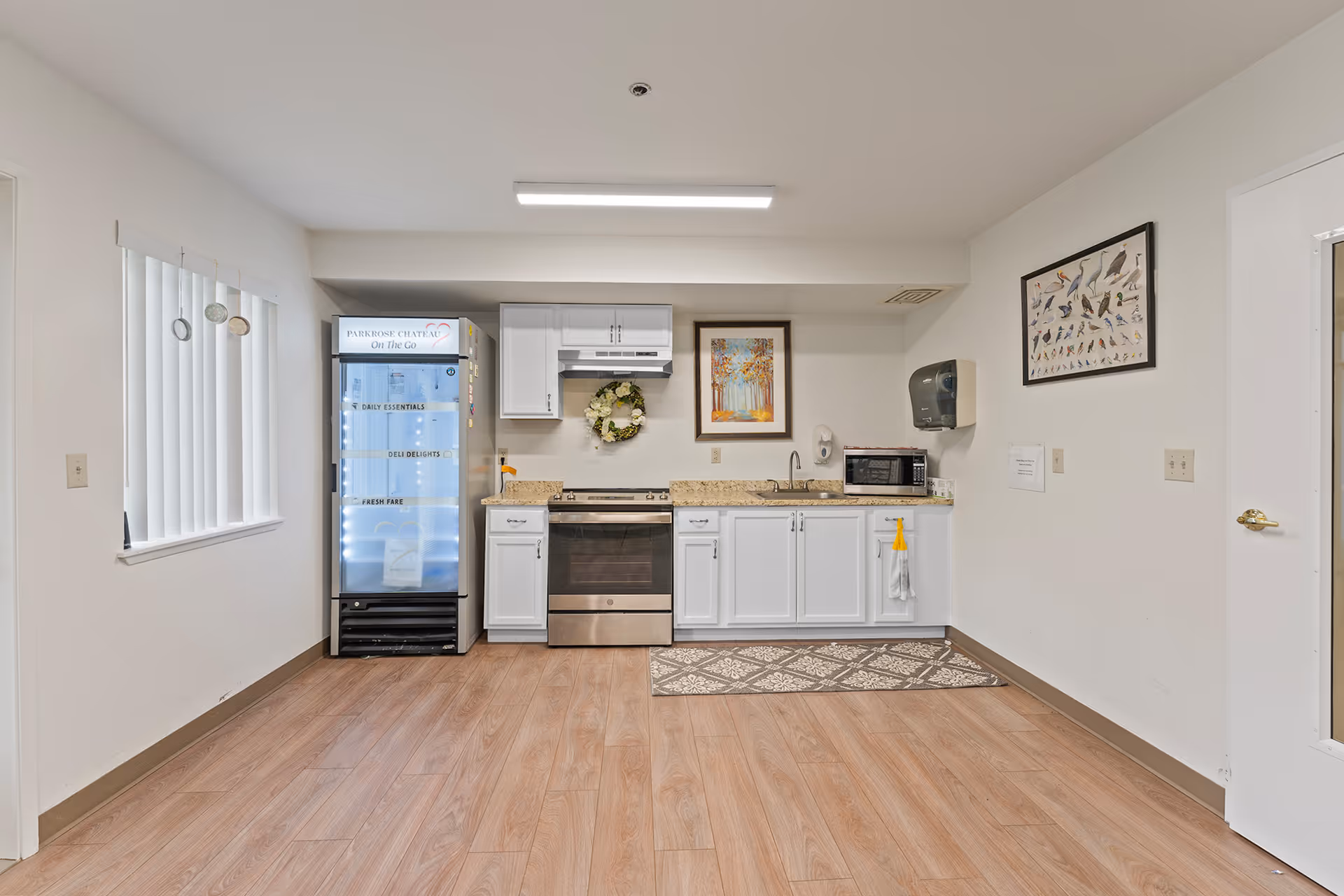 A clean and simple kitchen area with white cabinets, a stainless steel stove, a microwave, and a small sink. There is a refrigerator labeled 'Parkrose Chateau On The Go' with sections for daily essentials, deli delights, and fresh fare. The floor is wood laminate, and the walls are white with two framed pictures hanging. A paper towel dispenser and soap dispenser are mounted on the wall.