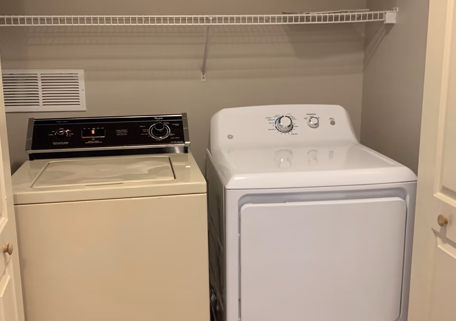 Laundry area with a beige top-loading washing machine on the left and a white front-loading dryer on the right, with a wire shelf above them and beige walls surrounding the machines.