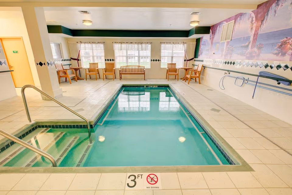 Indoor swimming pool area with clear water, metal handrails, and steps leading into the pool. Surrounding the pool are tiled floors, wooden chairs, and benches along the walls. Large windows with sheer curtains allow natural light to fill the room. A mural depicting an outdoor scene with arches and trees decorates one wall.
