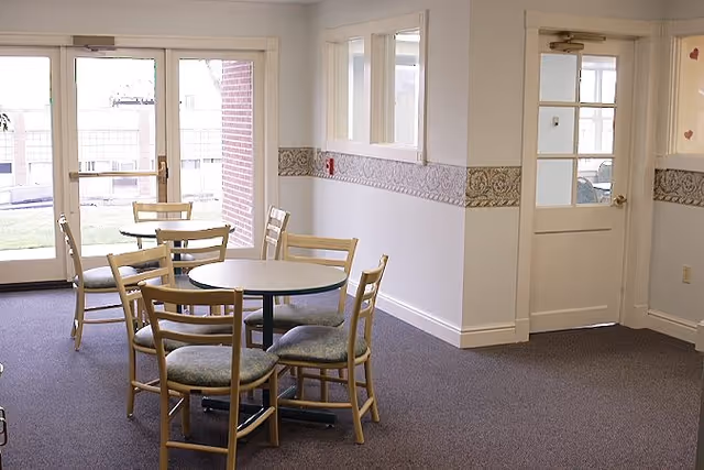 A small dining area with two round tables and several wooden chairs with padded seats. The room has carpeted flooring, white walls with a decorative border, and large glass doors leading outside to a grassy area.