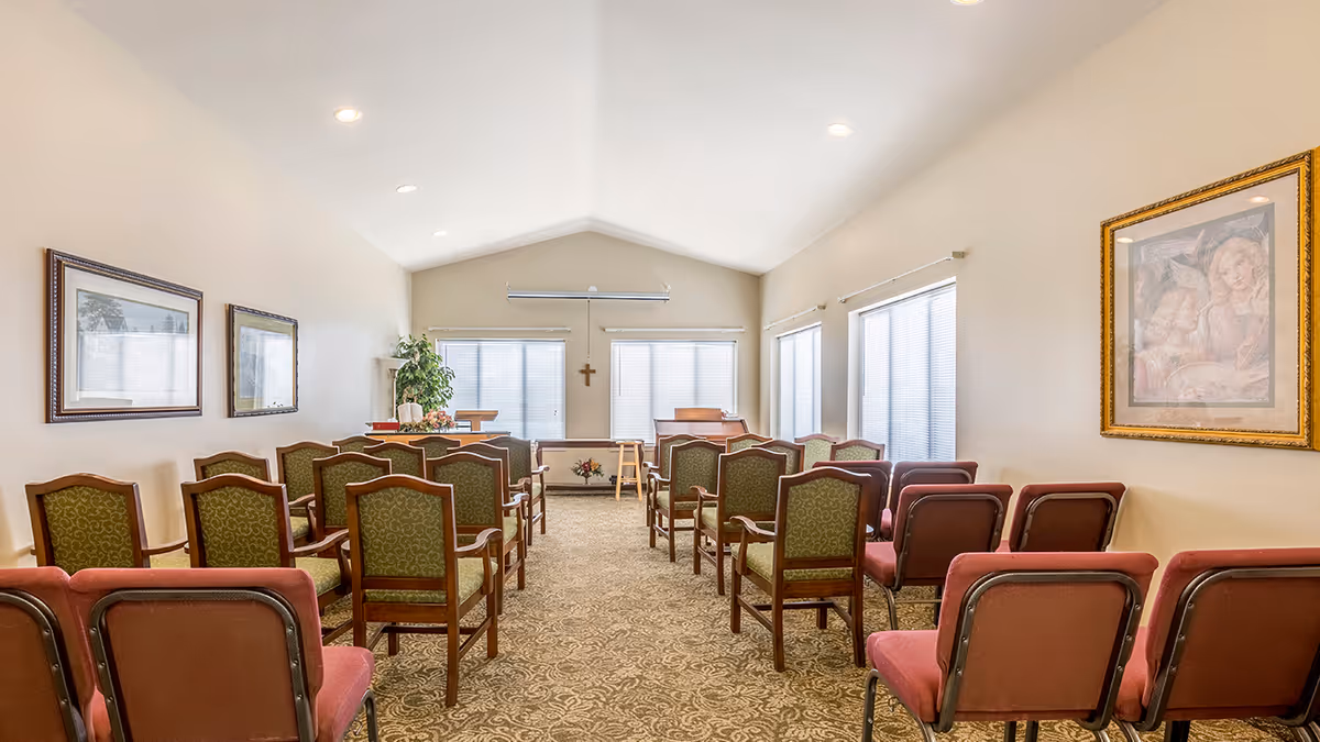 A small chapel or meeting room with rows of green and red chairs facing a podium and a cross on the wall. The room has beige walls, carpeted floor, framed pictures on the walls, and several windows with blinds.