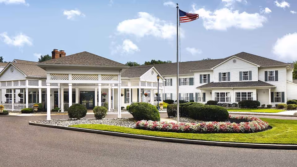 Front exterior view of The Residence at Voorhees, a large white building with multiple windows, a covered entrance, an American flag on a flagpole, and landscaped bushes and flowers in front.