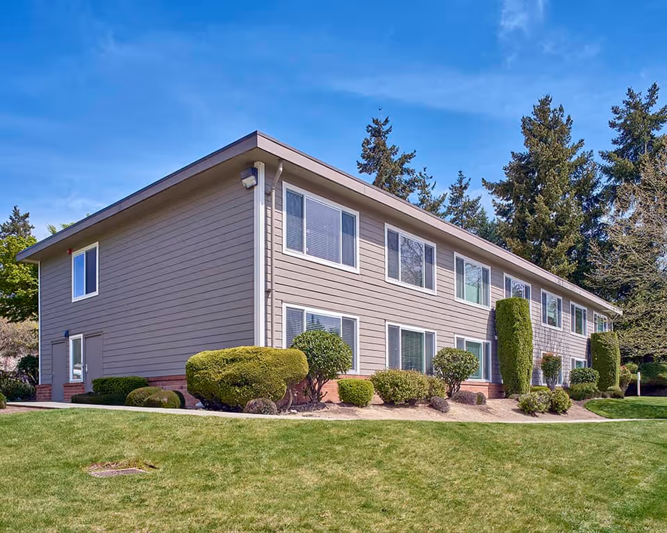 Exterior view of a two-story senior living facility building with beige siding and multiple windows, surrounded by neatly trimmed bushes and green grass under a clear blue sky.