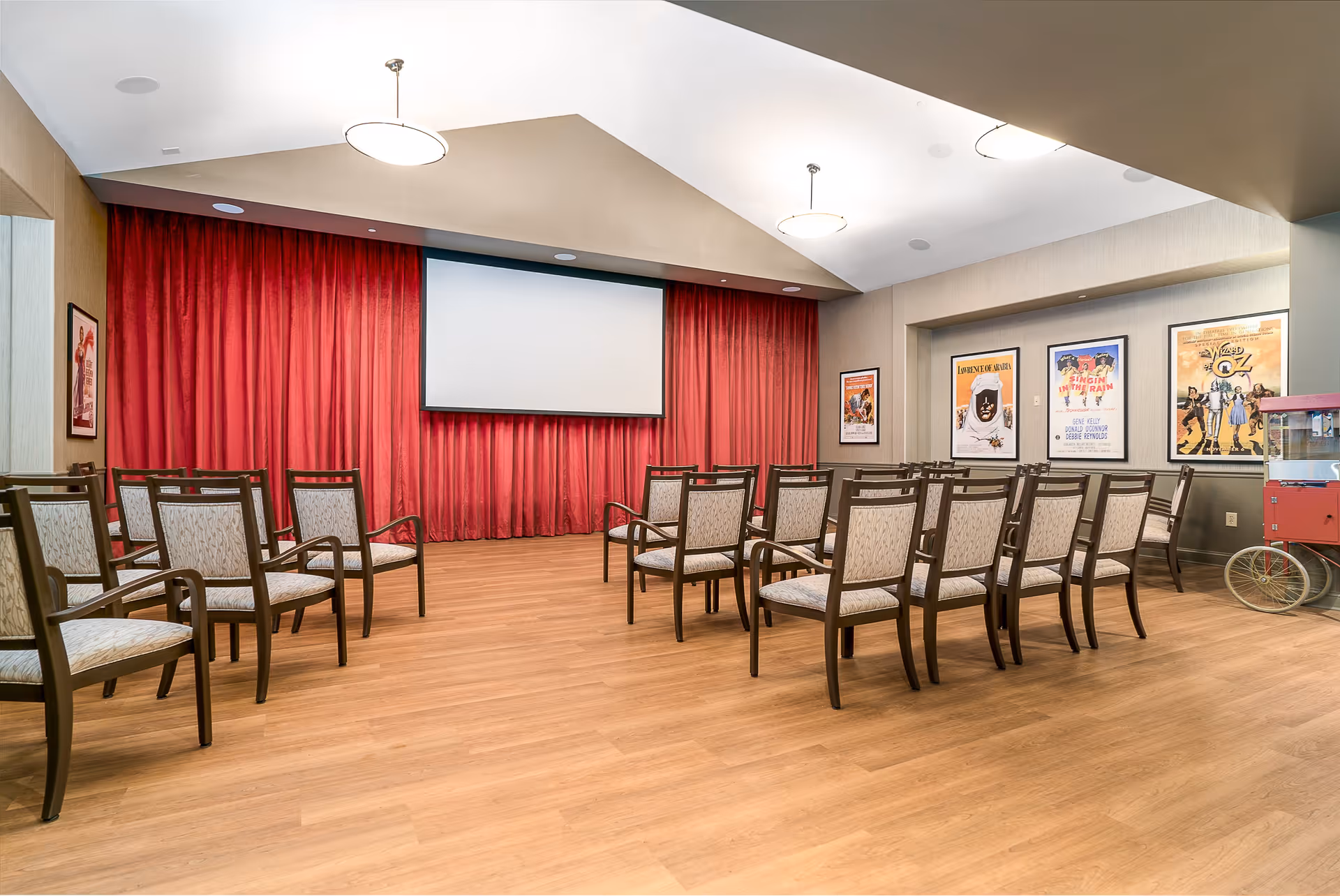 A small theater room with rows of chairs facing a large projection screen with red curtains behind it. The room has wooden flooring, beige walls, and movie posters on the walls. There is also a popcorn machine on the right side of the room.