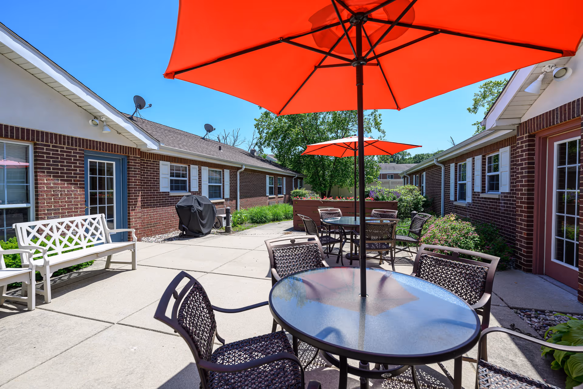 Outdoor patio area with round glass tables and metal chairs under large red umbrellas. The patio is surrounded by brick buildings with windows and doors, and there is a white bench and a covered grill visible. Green shrubs and trees are in the background under a clear blue sky.