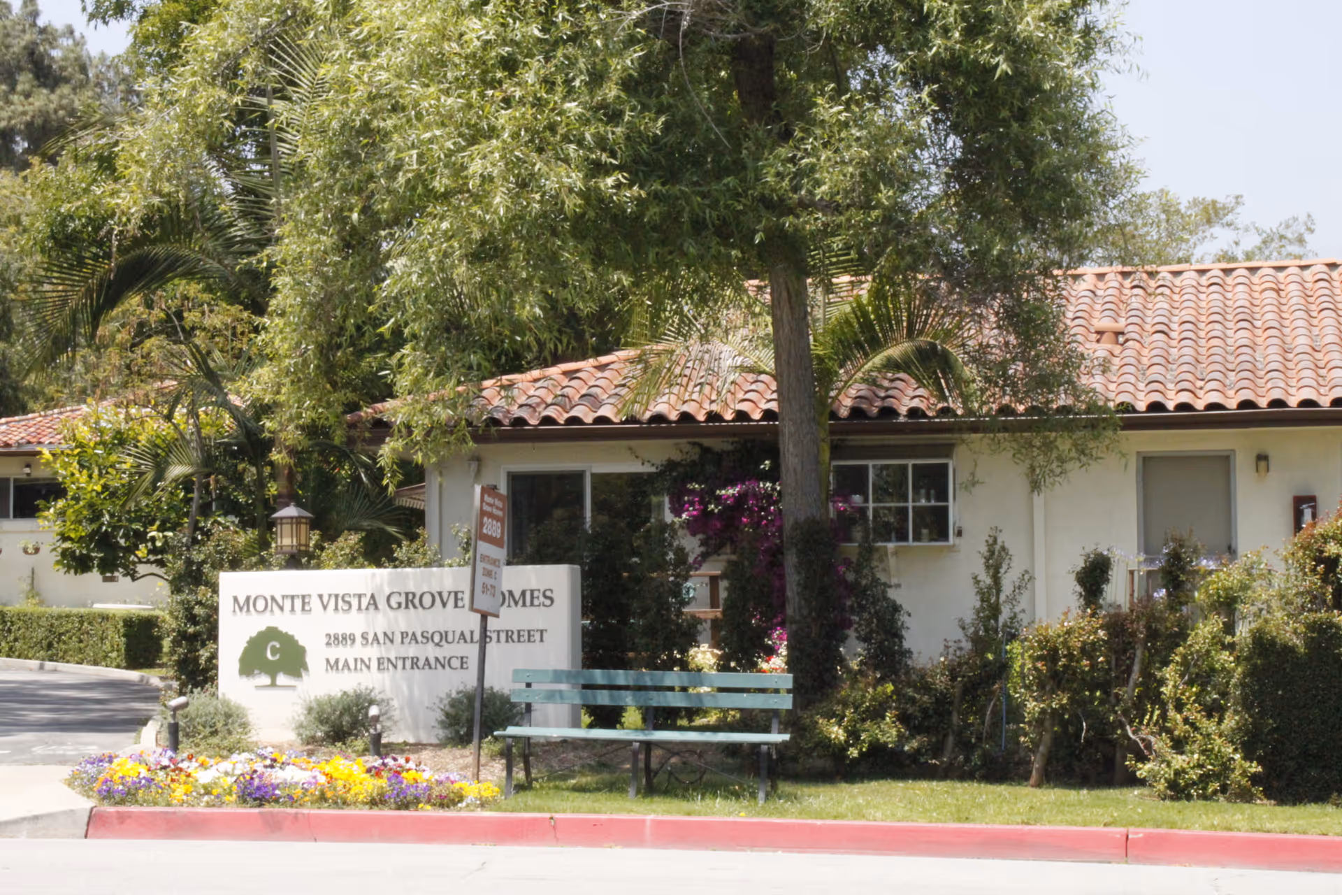 Exterior view of Monte Vista Grove Homes featuring a white building with a red tile roof, surrounded by trees and bushes. A sign in front reads 'Monte Vista Grove Homes, 2889 San Pasqual Street, Main Entrance.' There is a green bench and colorful flowers near the sign.