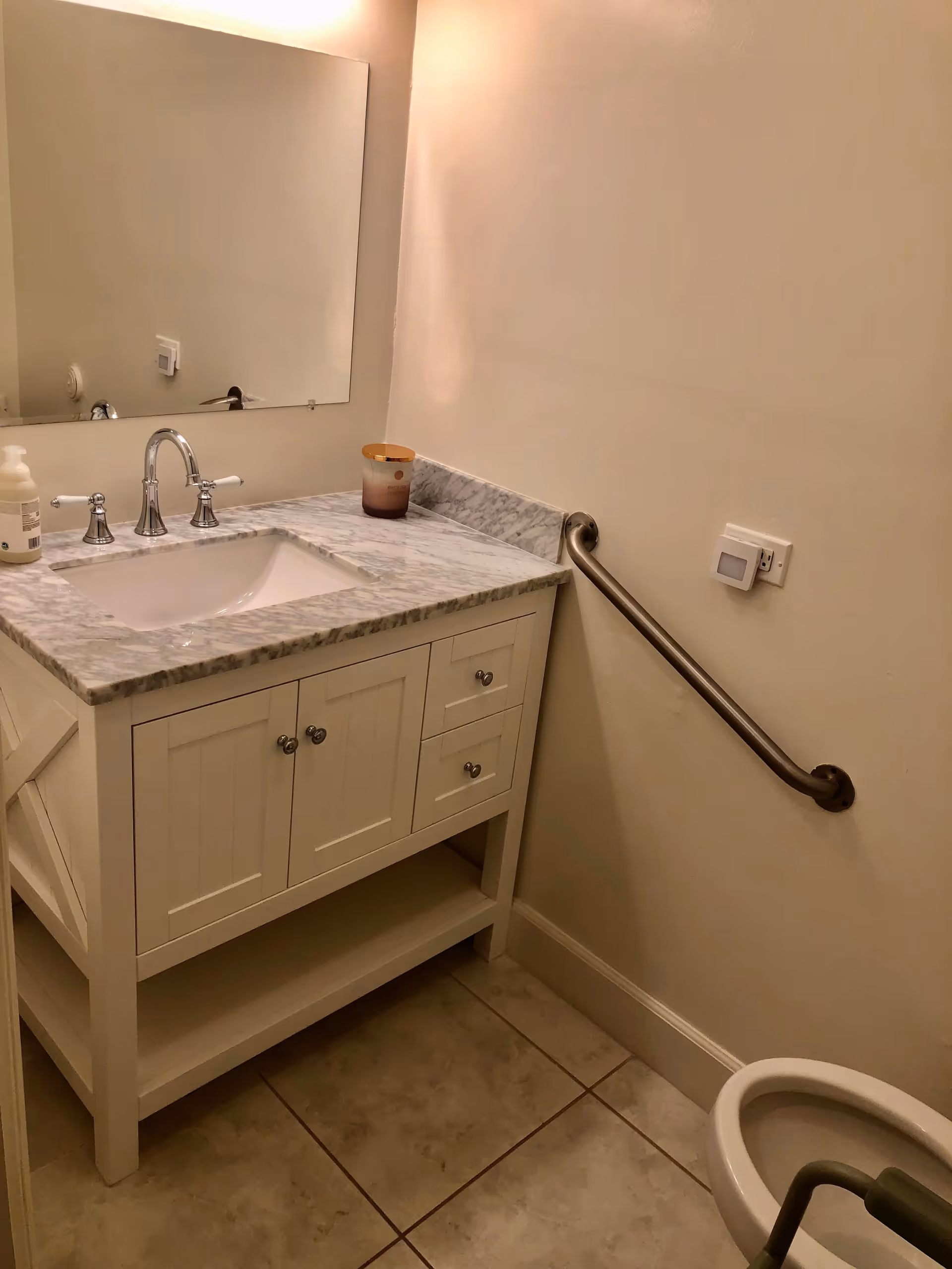 A small bathroom with a white vanity featuring a marble countertop and a built-in sink. There is a large rectangular mirror above the vanity. On the countertop, there is a soap dispenser and a candle. A stainless steel grab bar is mounted on the wall next to the vanity. The floor is tiled, and part of a toilet with a grab bar is visible in the lower right corner.