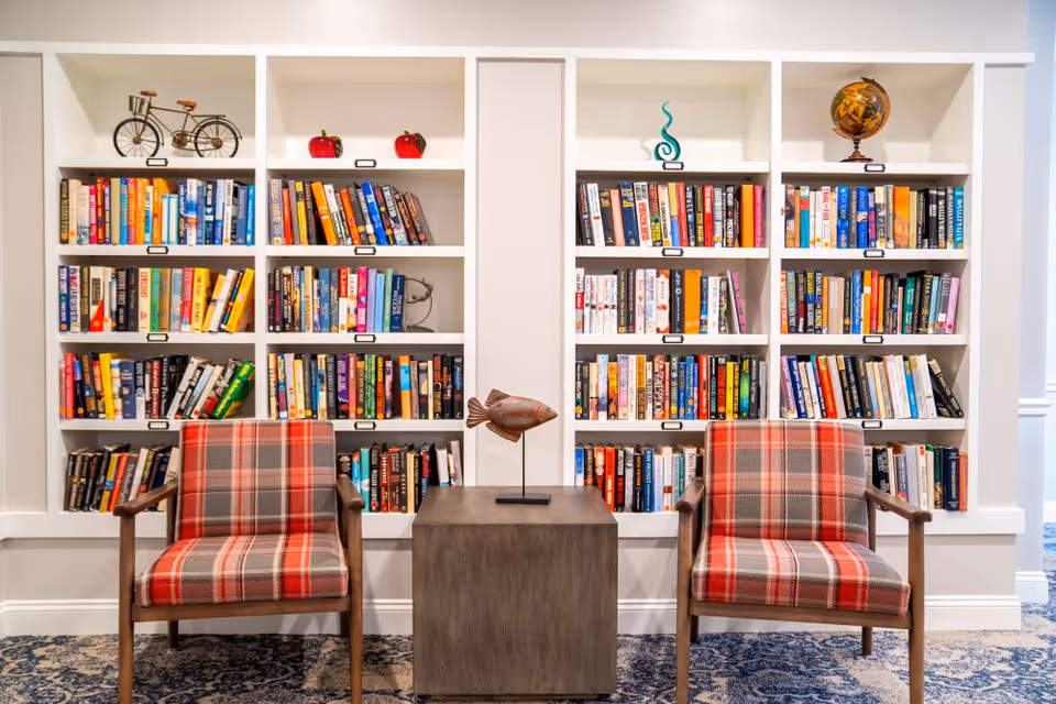 A cozy reading nook featuring two plaid upholstered armchairs with wooden arms, positioned on either side of a small square wooden table with a decorative fish sculpture. Behind the chairs is a white built-in bookshelf filled with a variety of colorful books and decorative items including a small bicycle model, two red apple ornaments, a blue abstract sculpture, and a globe. The area has a patterned carpet and white walls.