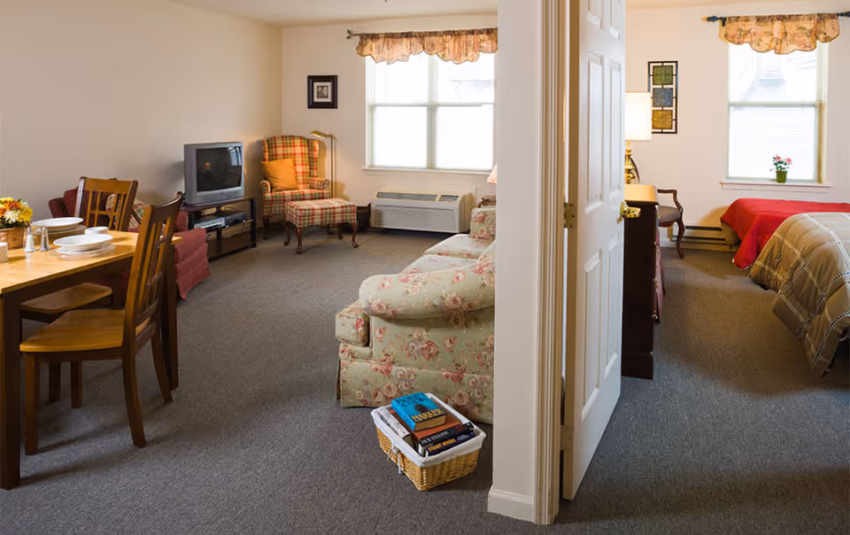 Interior of a senior living apartment showing a living room with a floral sofa, armchair, TV and dining table, with a bedroom visible through an open door.