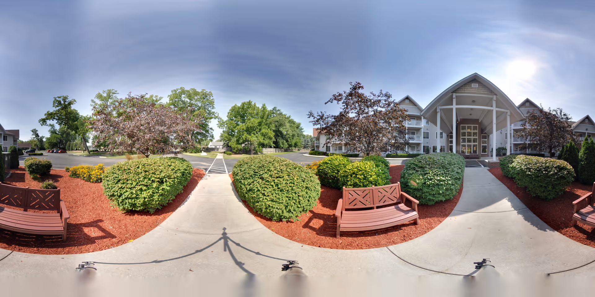 Outdoor view of the entrance to Charter Senior Living of Kenosha, showing a paved walkway flanked by well-maintained bushes and red mulch beds, wooden benches on either side, and a large building with a covered entryway under a clear sky with the sun shining.