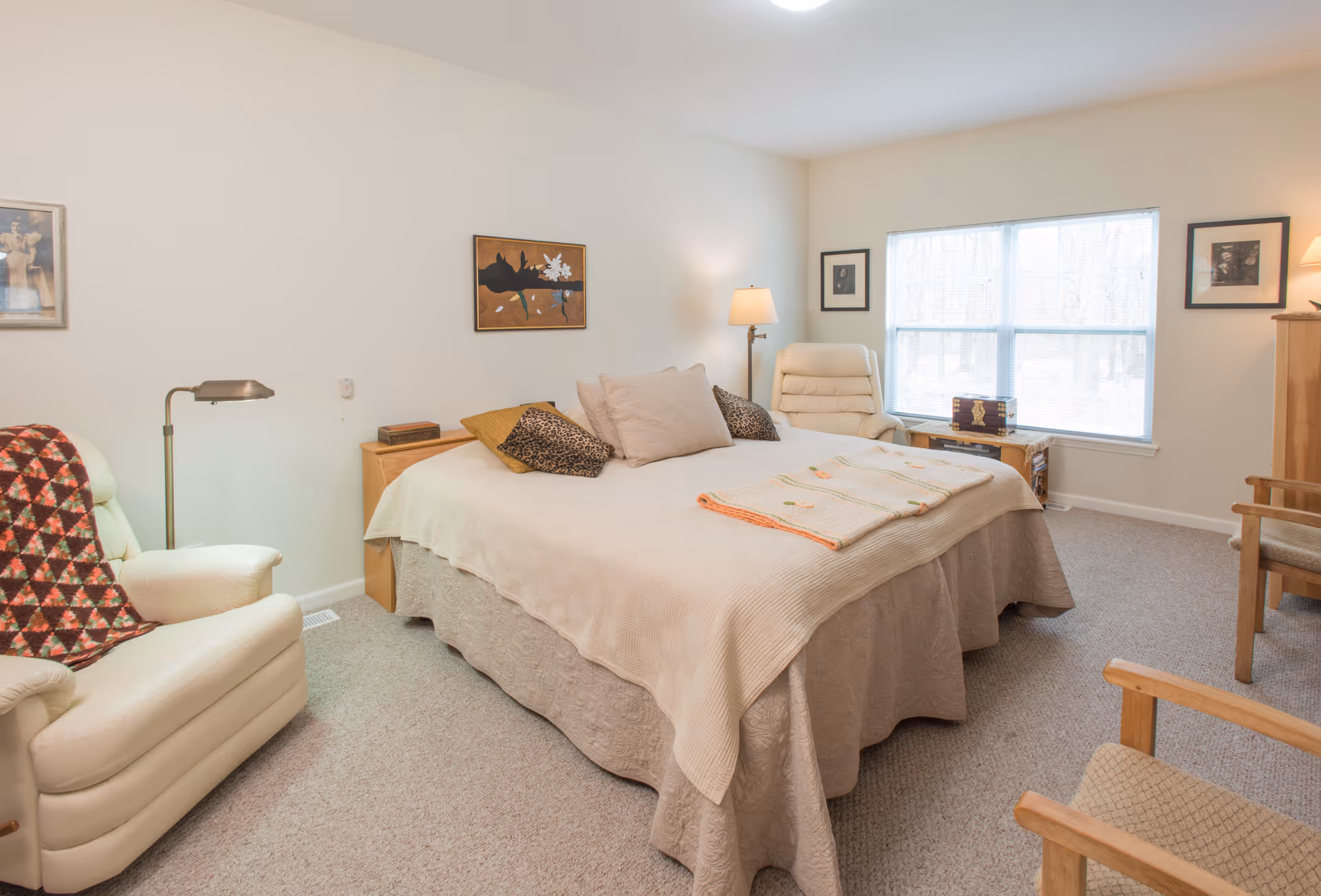 A cozy bedroom with a large bed covered in beige and light brown bedding, surrounded by several pillows. There are two armchairs, one with a patterned blanket draped over it, a floor lamp, and a wooden side table. The room has beige walls with framed artwork and a large window letting in natural light. The carpet is light-colored, and the overall atmosphere is warm and inviting.