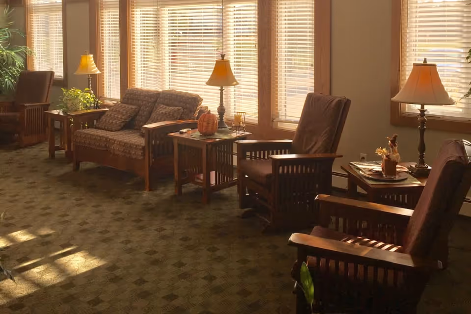 A cozy living room area with wooden framed armchairs and a loveseat, each with cushions. There are side tables with lamps and decorative items, and large windows with blinds letting in natural light. The room has a patterned carpet and some plants in the corners.