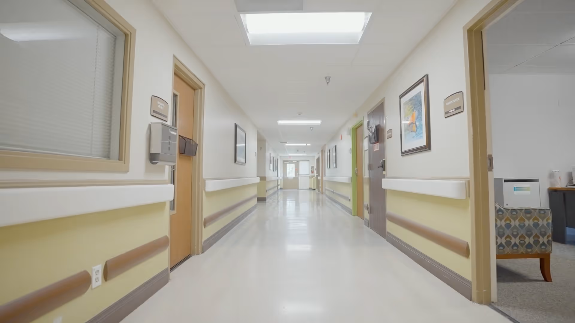 A clean, well-lit hallway in a senior living facility with beige and yellow walls, handrails on both sides, several closed doors, framed artwork on the walls, and a skylight providing natural light.