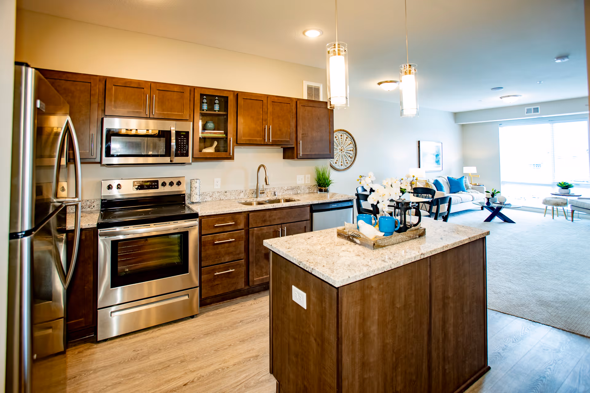 A modern kitchen with stainless steel appliances including a refrigerator, oven, and microwave. The kitchen features dark wooden cabinets, granite countertops, and a central island with decorative items. In the background, there is a living area with a sofa, chairs, and large windows letting in natural light.