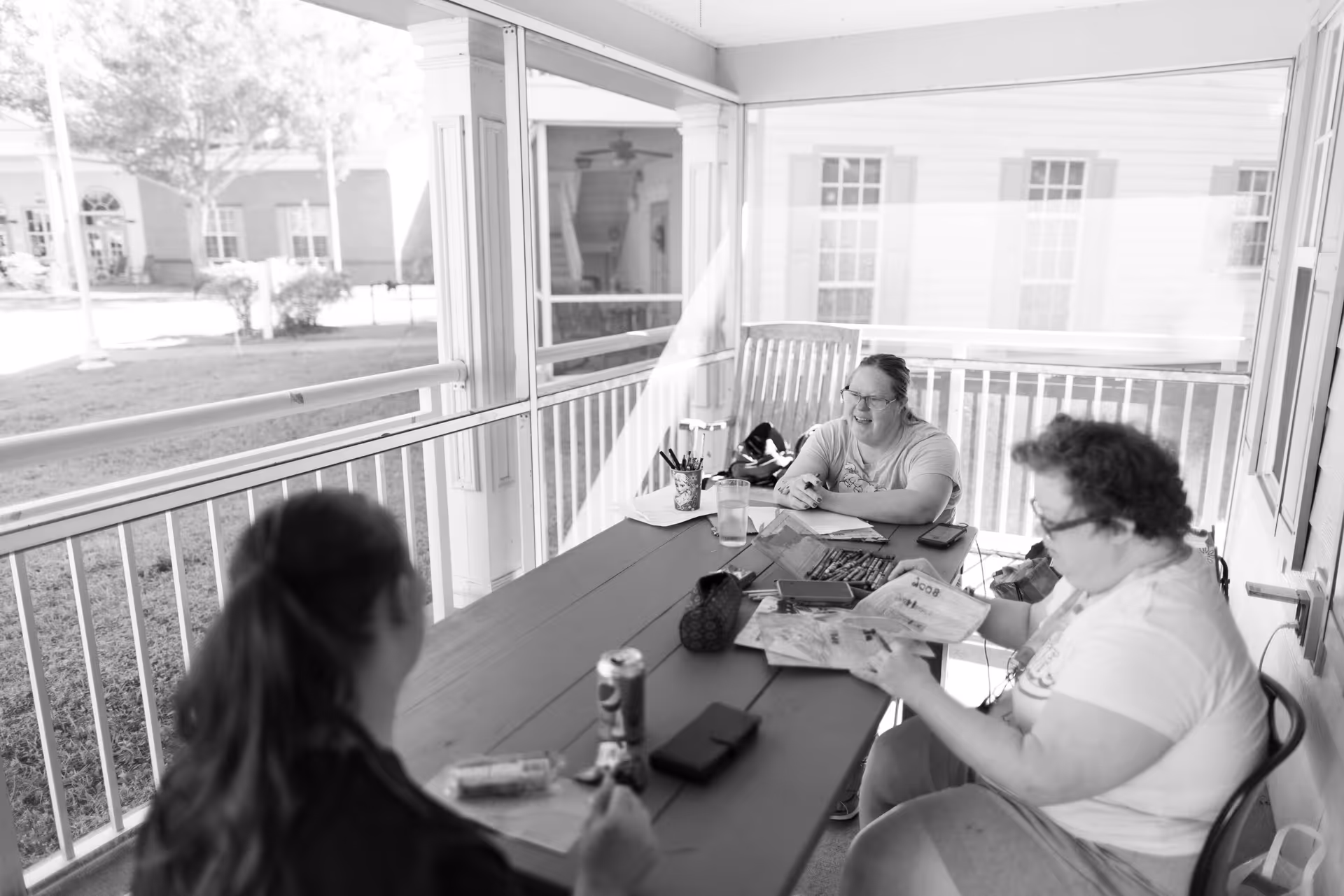Three people sitting around a table on a covered porch engaged in an activity with papers and coloring supplies, with a grassy outdoor area visible through the porch railing.