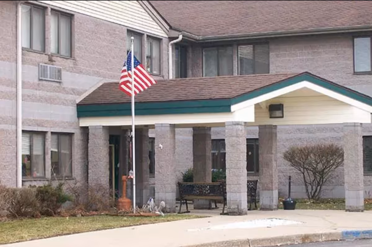 Exterior view of Wood Ridge Assisted Living facility showing the entrance with a covered porch supported by stone pillars, an American flag on a flagpole, benches, and some landscaping with bushes and grass.
