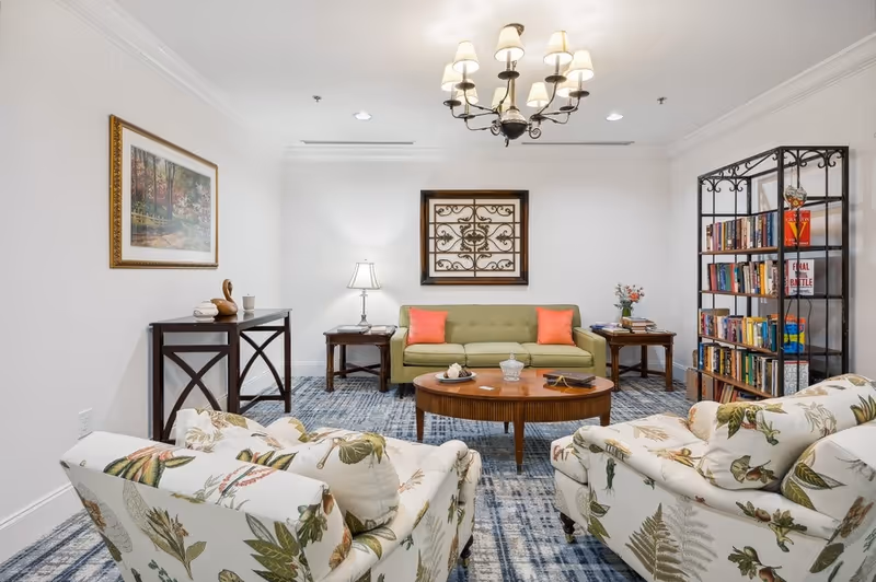 A cozy living room with a green sofa adorned with two coral pillows, two floral-patterned armchairs, a round wooden coffee table, a bookshelf filled with books, two side tables with lamps and flowers, a chandelier hanging from the ceiling, and framed artwork on the walls.