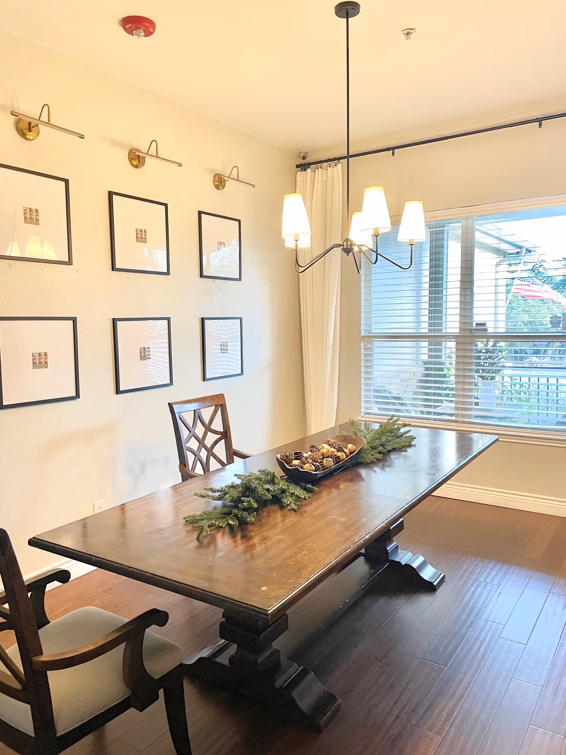 A dining room with a long wooden table decorated with a centerpiece of pine branches and pine cones. Two wooden chairs with cushioned seats are positioned at the table. The room has six framed pictures on the wall, a window with blinds, and a modern chandelier with five lights hanging from the ceiling.