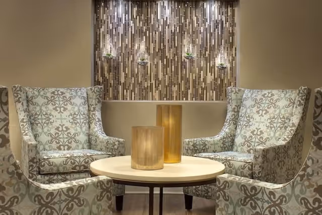 A seating area with patterned armchairs around a round table topped with gold candle holders, set before a mosaic tiled accent wall with small hanging plants.