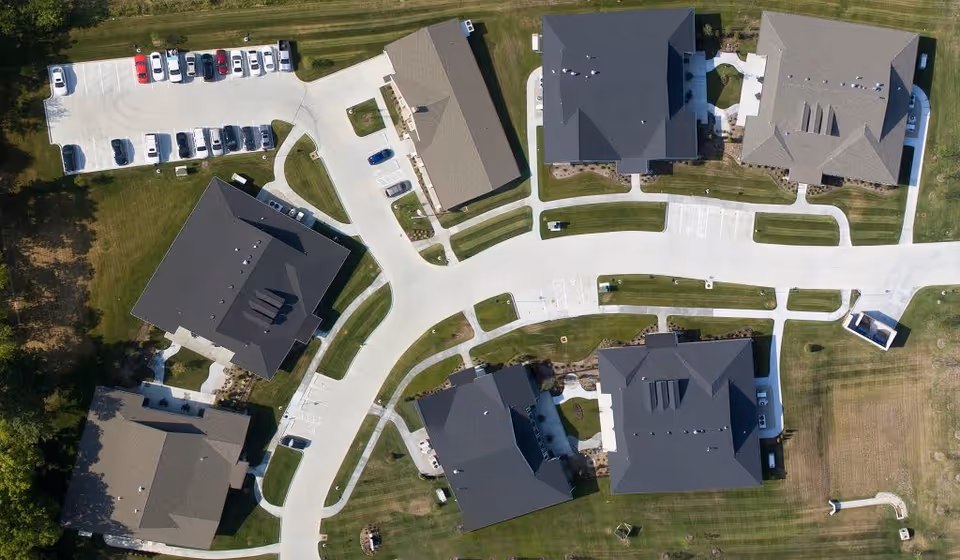 Aerial view of the Cottages of Lake St Louis senior living facility showing multiple residential buildings with dark roofs, paved roads and parking areas, and well-maintained green lawns surrounding the buildings.