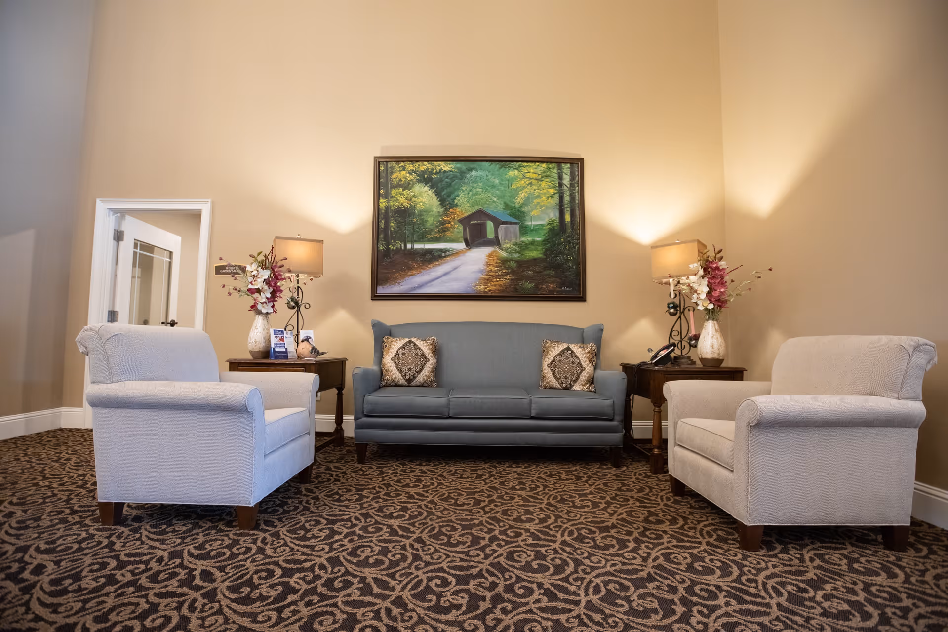 Lobby-style seating area with a blue sofa, two armchairs, side tables, lamps and a landscape painting on the wall.