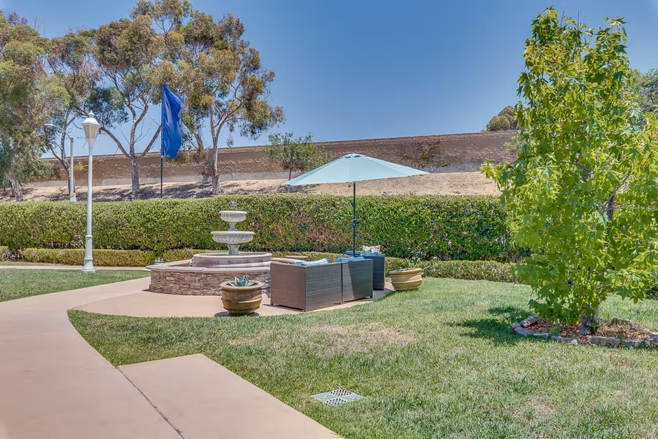 Outdoor seating area with a wicker sofa and chairs under a blue umbrella next to a stone water fountain, surrounded by green grass, trees, and a hedge with a clear blue sky in the background.