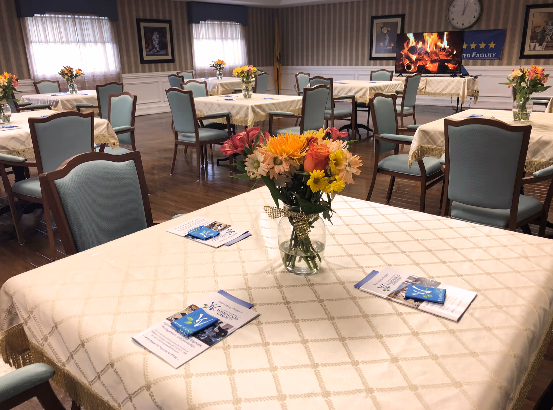 A dining room with multiple tables covered in cream-colored tablecloths, each adorned with a vase of colorful flowers. The chairs around the tables have light blue upholstery and wooden frames. In the background, there is a TV screen displaying a fireplace scene, a clock on the wall, and framed artwork. The room has wooden flooring and windows with sheer curtains.