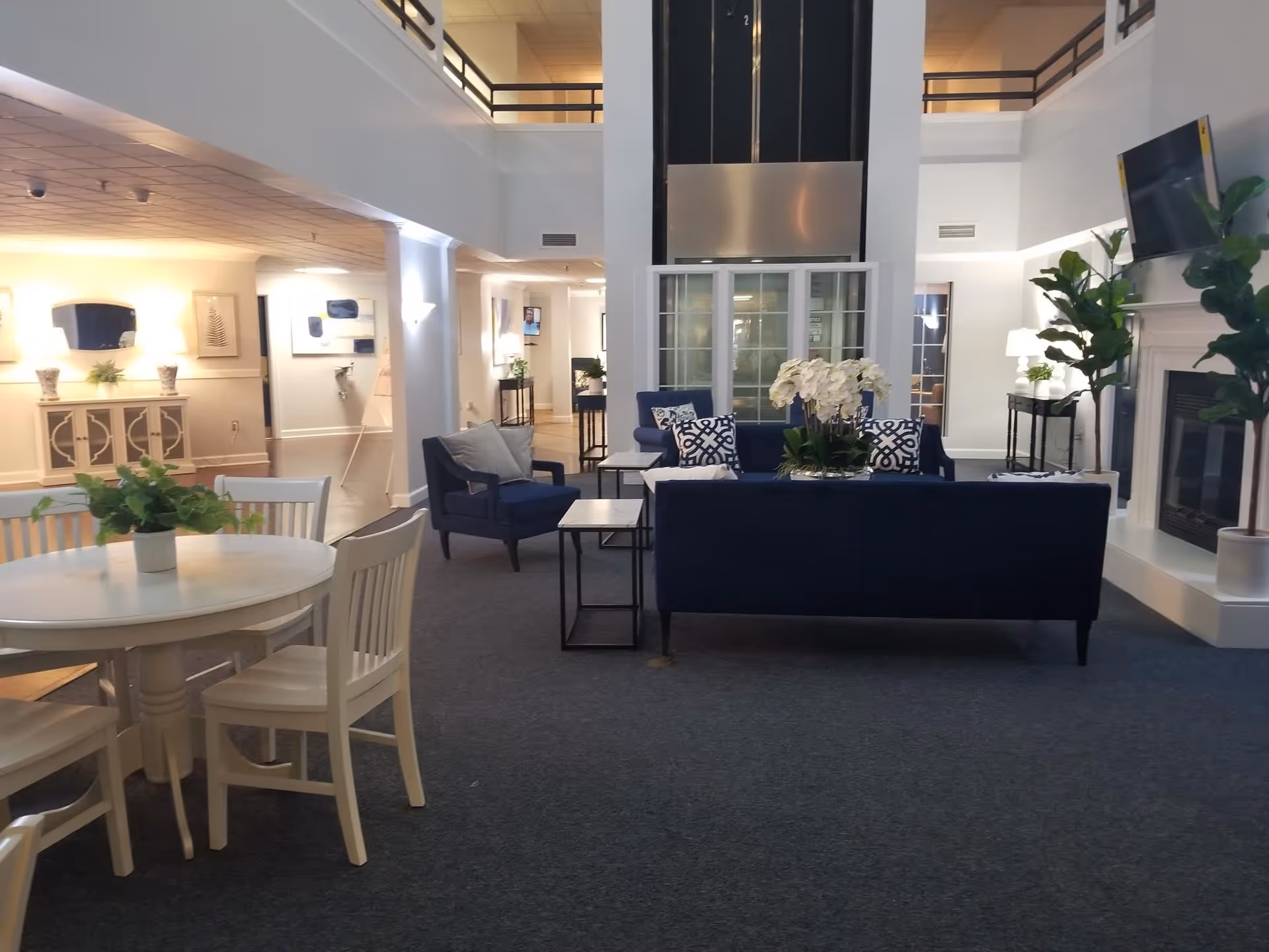 A spacious and well-lit senior living facility common area with a seating arrangement including a navy blue sofa and armchairs around a coffee table with white flowers. There is a round dining table with white chairs and a green plant in the foreground. The room features a fireplace with a mounted TV above it, potted plants, and decorative wall art. The ceiling is high with a second-floor balcony visible.