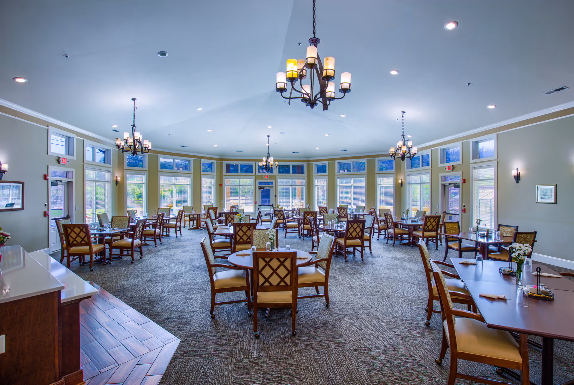 Spacious, well-lit dining room with multiple round and rectangular tables and chairs, chandeliers, and large windows.