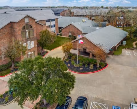 Aerial view of a senior living facility named Serenity at Briarcrest showing multiple brick buildings with gray roofs, surrounded by trees and landscaping. There is a parking lot with several cars and a flagpole with two flags near the entrance.