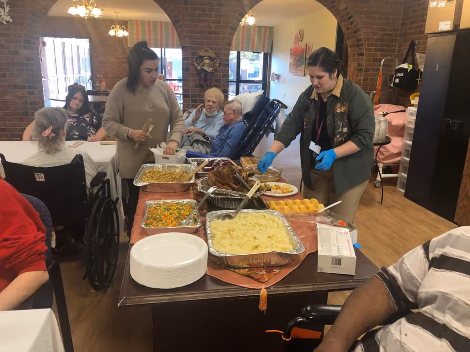 Staff members serving a buffet-style meal to elderly residents seated at tables in a brick-walled dining room.