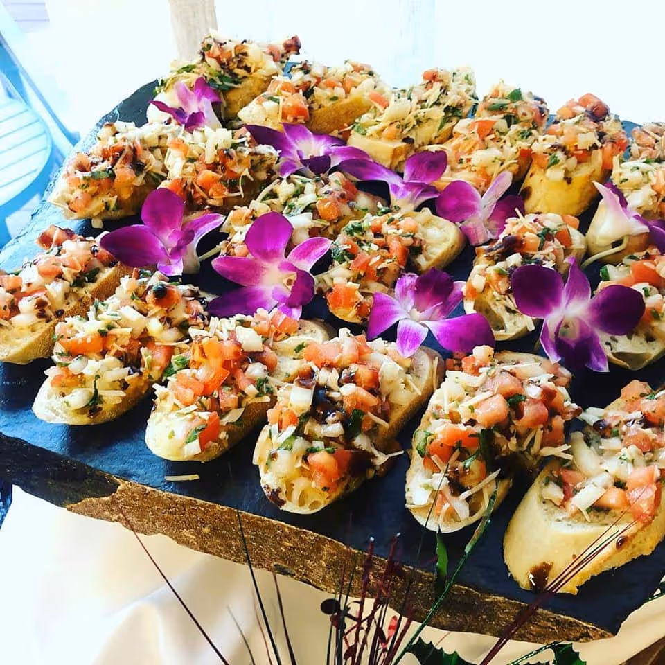 A platter of bruschetta topped with diced tomatoes, herbs, and shredded cheese, garnished with purple orchid flowers, displayed on a rustic wooden serving board.