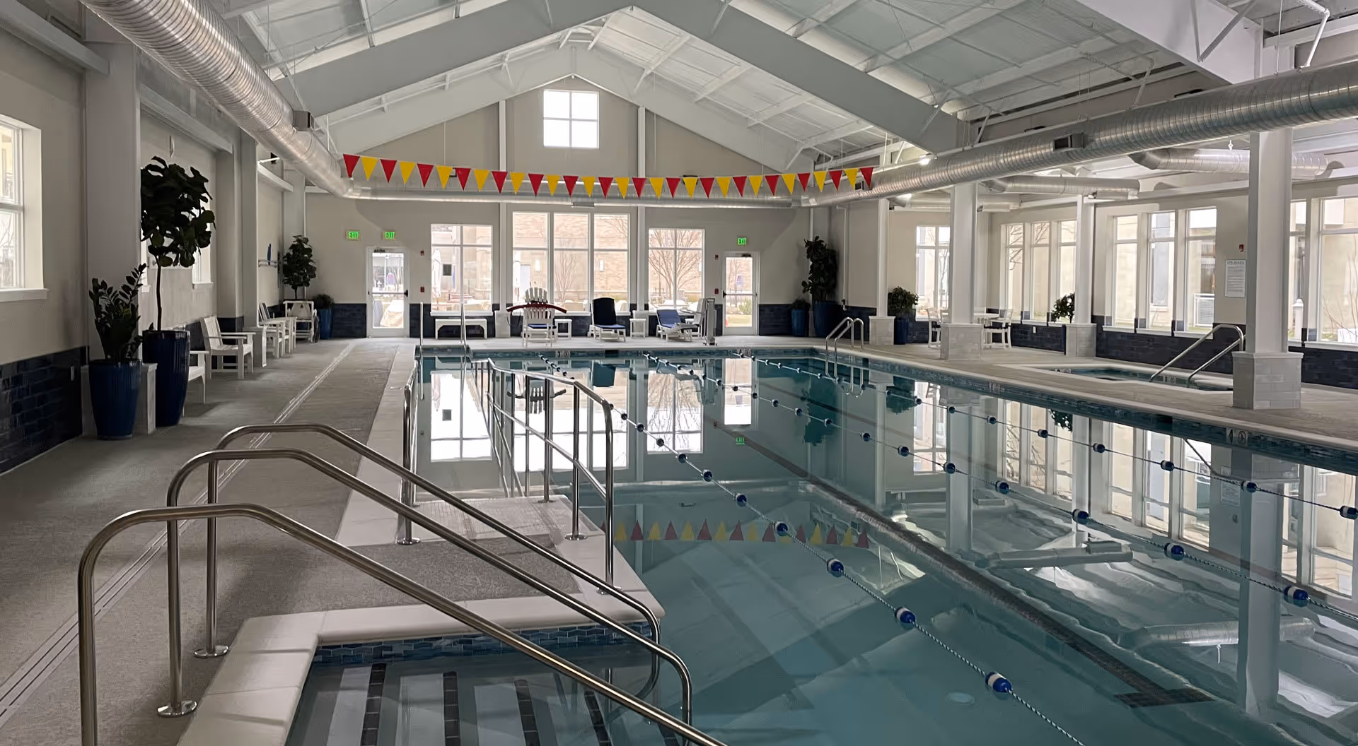 Indoor swimming pool area with clear water, stainless steel handrails, and lane dividers. The space has large windows letting in natural light, potted plants along the walls, white benches, and a high ceiling with exposed ductwork. Colorful triangular flags hang across the room near the ceiling.