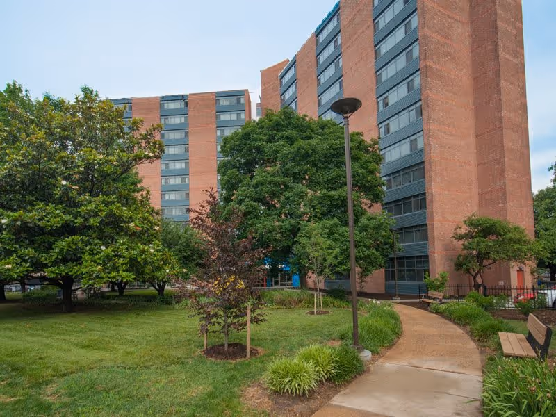 Outdoor view of Linden Park Apartments in Bolton Hill showing tall brick residential buildings surrounded by green trees, grass, and a paved walkway with benches and a streetlamp.