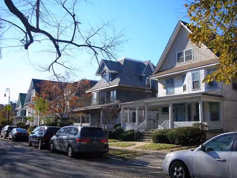 Row of residential-style buildings with porches along a tree-lined street and parked cars.