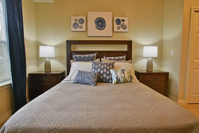 Well-lit bedroom with a neatly made bed, decorative pillows, matching nightstands with lamps, and artwork above the headboard.