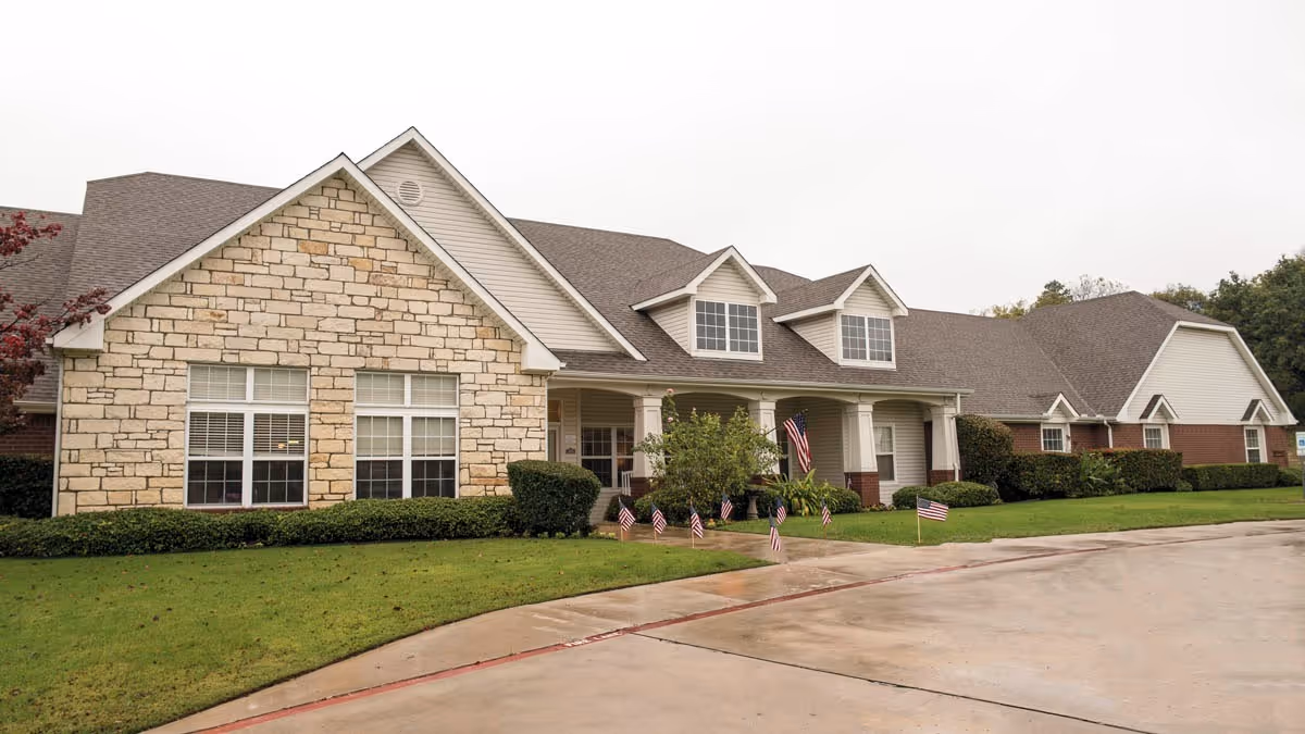 Exterior view of a single-story senior living facility building with a stone and brick facade, multiple windows, a covered entrance with columns, small American flags placed along the front lawn, and a paved driveway in front.