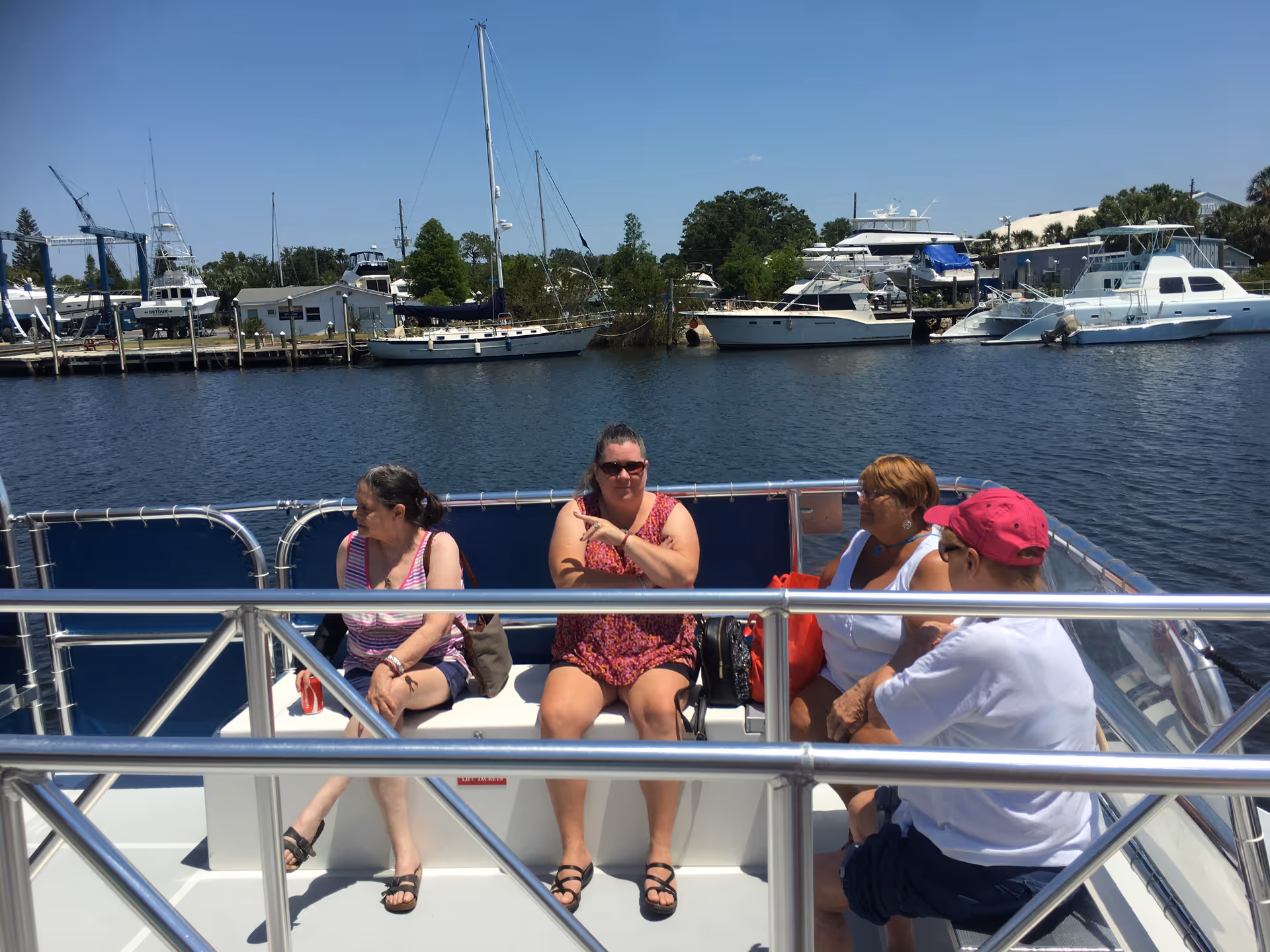 Four women sitting and chatting on a boat with a marina and several docked boats in the background under a clear blue sky.