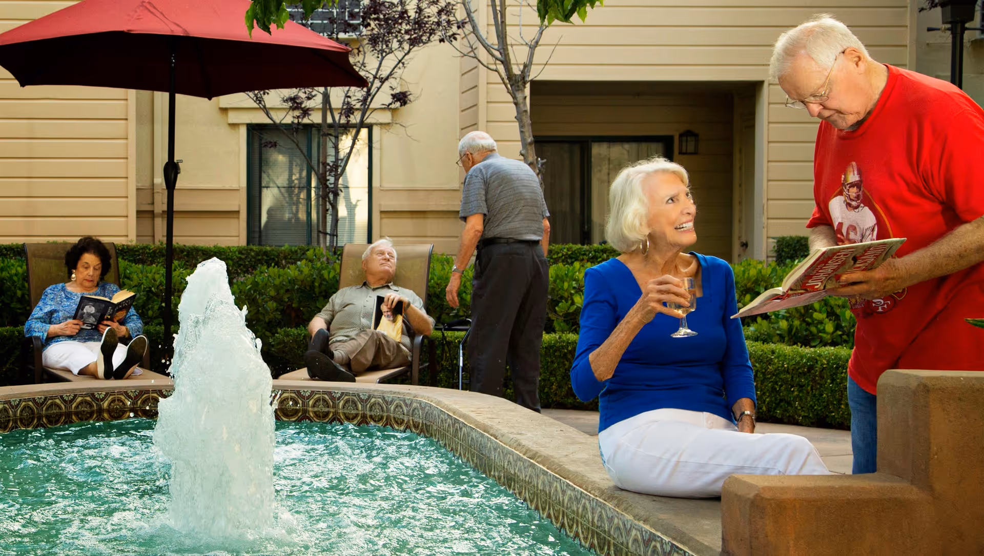 An outdoor courtyard at Chateau Cupertino featuring a water fountain in the center. Five elderly people are enjoying the space: one woman in a blue top and white pants is sitting on the edge of the fountain holding a drink and smiling at a man in a red shirt who is showing her a book. Two other people are seated on lounge chairs reading, and another man is standing near the bushes. The area is surrounded by greenery and beige building walls.