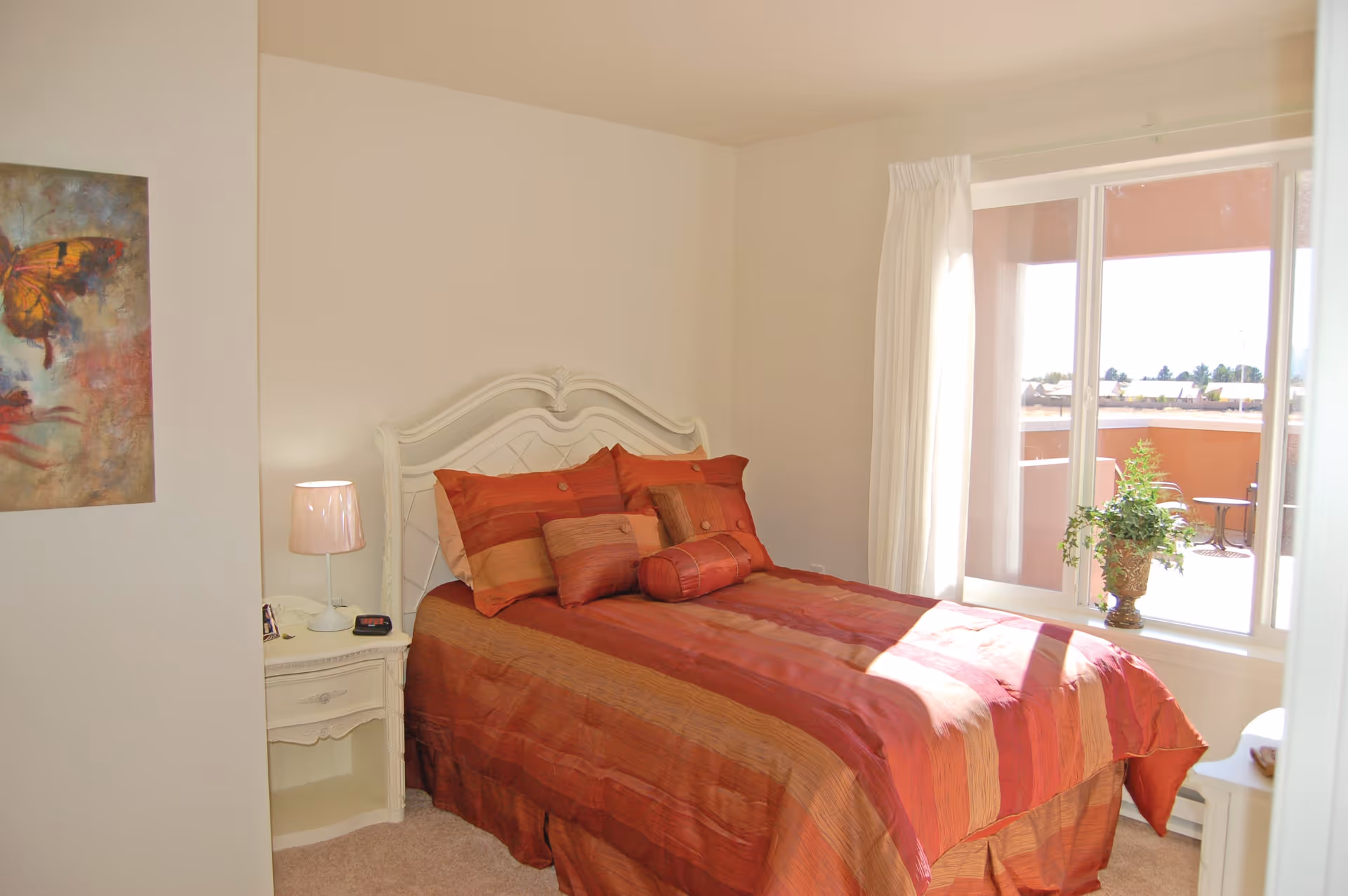 A cozy bedroom with a bed dressed in red and orange striped bedding, a white ornate headboard, a nightstand with a lamp and clock, a colorful butterfly painting on the wall, and a large window with white curtains letting in natural light and showing a balcony with outdoor furniture and plants.