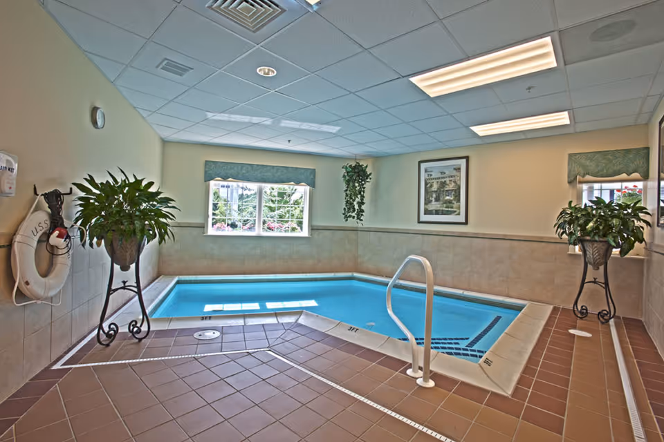 Indoor small swimming pool with tiled floor and walls, two large potted plants on decorative stands, a life preserver mounted on the wall, windows with valances letting in natural light, and framed artwork on the walls.