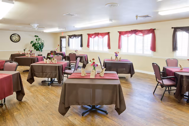 A bright dining room with several tables covered in brown tablecloths and red runners, each set with folded napkins and small flower arrangements. The room has wooden flooring, multiple windows with red and brown curtains, a wall clock, and a potted plant near the back door.