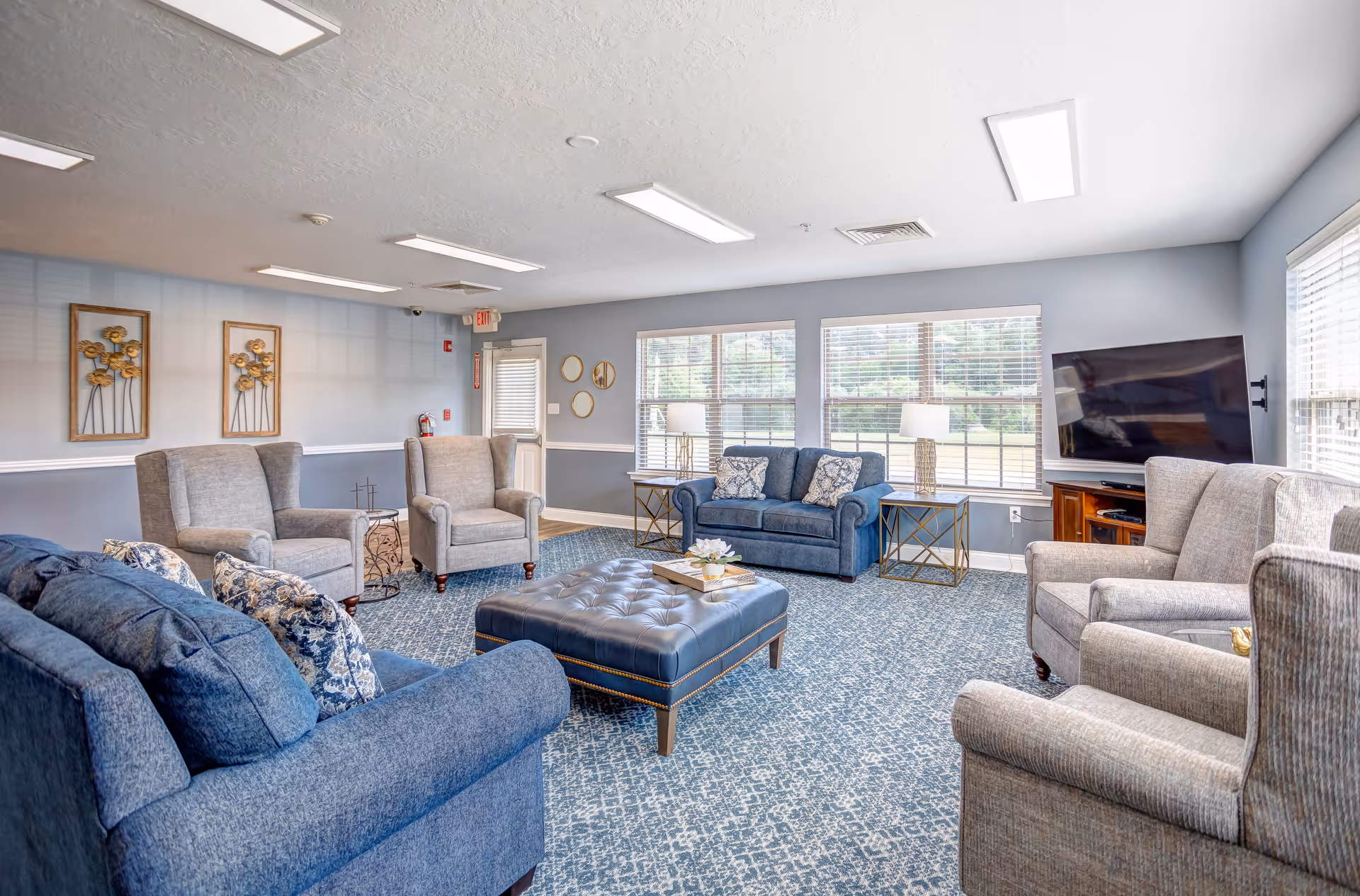 A bright and spacious living room with multiple upholstered armchairs and sofas arranged around a large tufted ottoman. The room features large windows with blinds, two side tables with lamps, a flat-screen TV mounted on the wall, and decorative wall art. The carpet has a blue patterned design, and the walls are painted light gray with white trim.