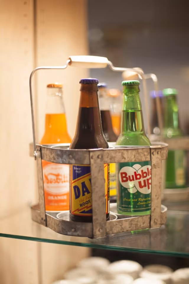 A metal carrier holding six glass soda bottles with various labels including root beer, orange soda, and Bubble Up, placed on a glass shelf.