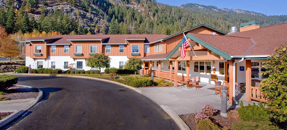 Exterior view of Mountain Meadows Senior Living Campus showing a two-story building with a brown roof and light brown and white walls, surrounded by landscaped bushes and trees, with mountains and pine trees in the background. There is a covered entrance with wooden posts, an American flag, and benches on the sidewalk.