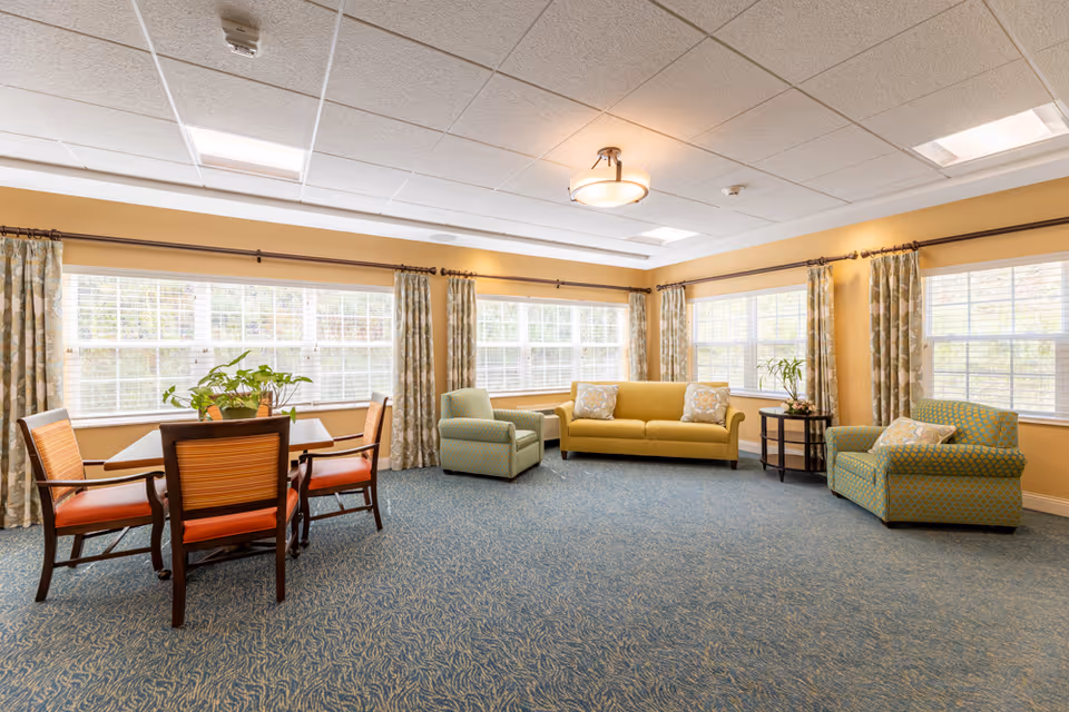 A bright and spacious living room with large windows covered by patterned curtains. The room features a yellow sofa with two patterned pillows, two green armchairs with patterned cushions, a wooden side table with a plant, and a wooden dining table with four chairs. The walls are painted a warm yellow, and the ceiling has recessed lighting and a central light fixture.