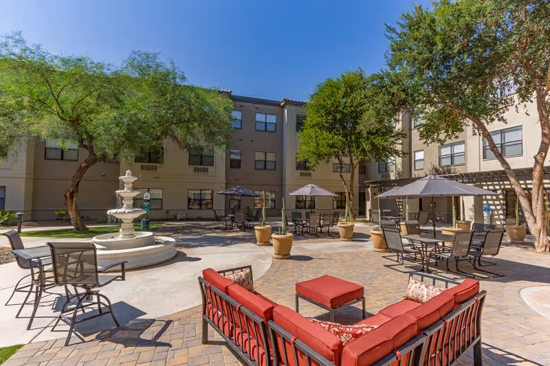 Outdoor courtyard area of a senior living facility with a multi-tiered white fountain, several seating areas including a red cushioned sectional sofa with matching ottoman, multiple tables with chairs and umbrellas, and trees providing shade. The building surrounds the courtyard with multiple windows visible under a clear blue sky.