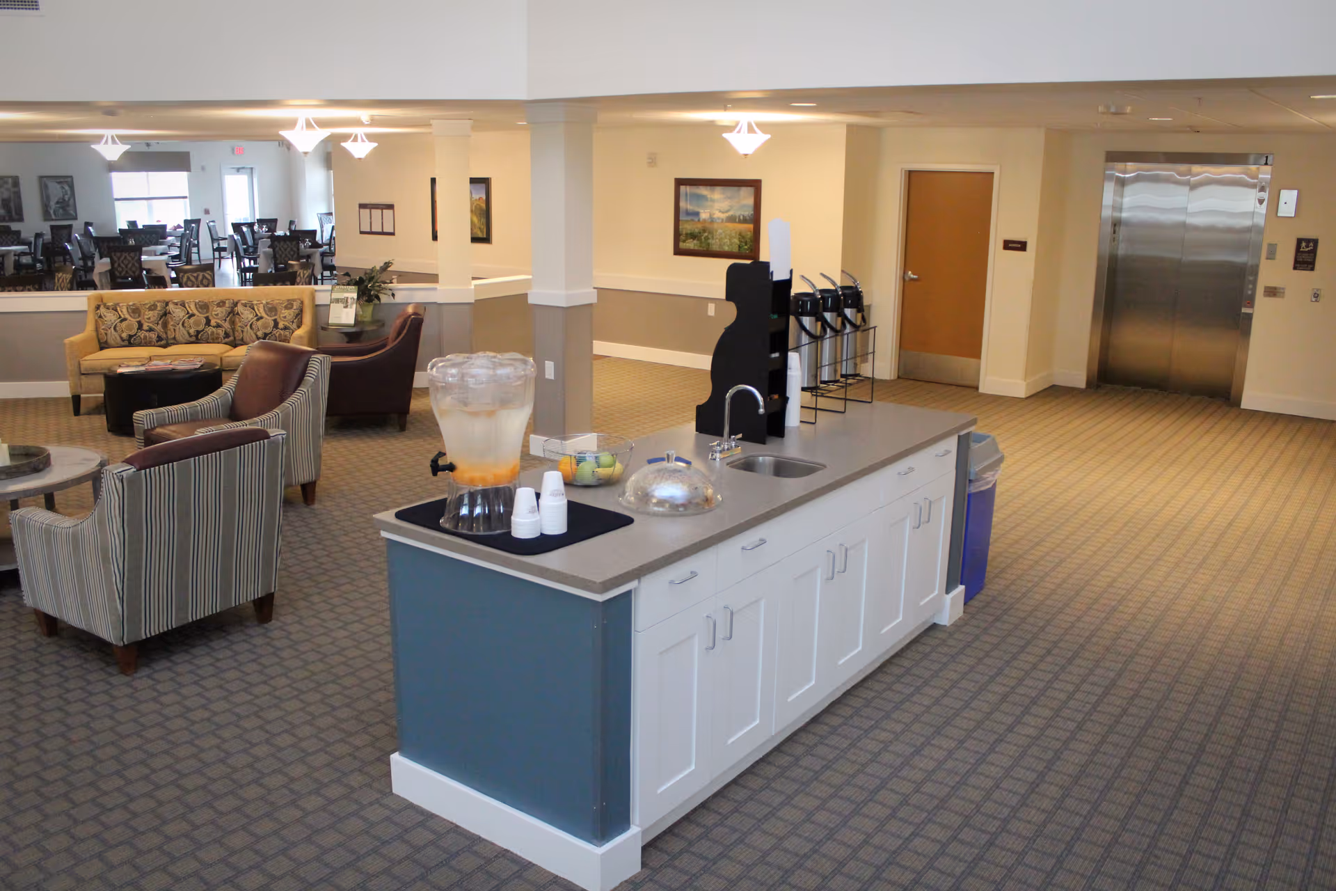 Open common area in a senior living facility with a central beverage island, lounge seating, and dining tables in the background.
