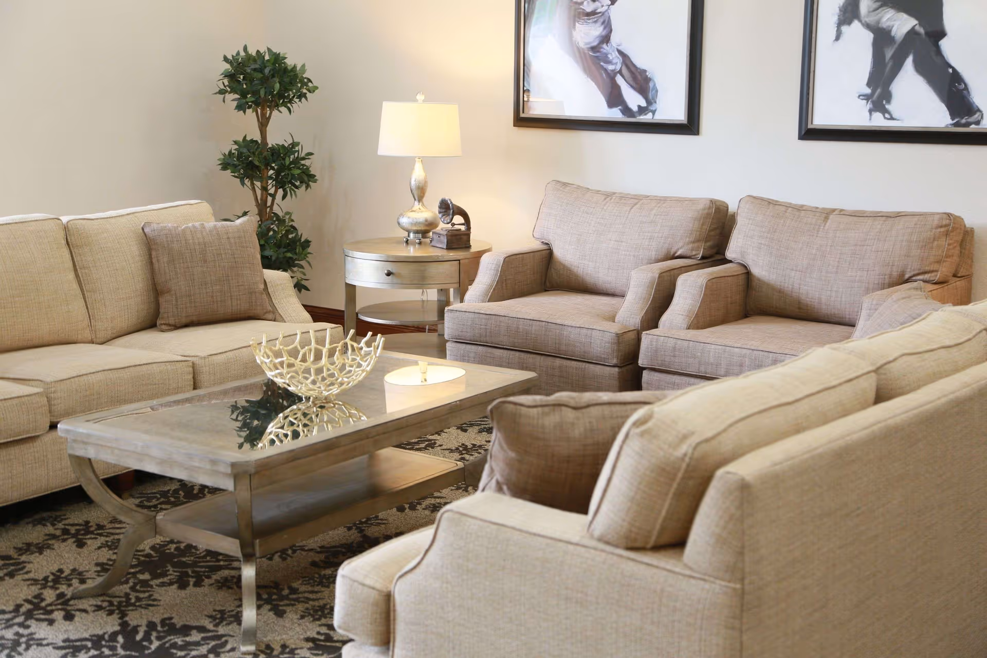 A cozy living room with beige sofas and armchairs arranged around a glass-top coffee table. A round side table with a lamp and decorative item is placed against the wall, with two framed black and white pictures hanging above. A green potted plant is in the corner, and a patterned rug covers the floor.